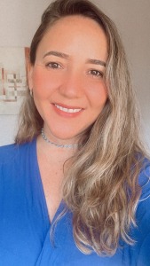 Foto da servidora Lucíola. Trata-se de uma mulher branca, com cabelo longo e loiro. Está vestindo uma camiseta azul e um colar prateado. Ela sorri para a foto.