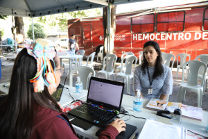 Foto da servidora Ana Flavia durante cadastro no sistema para doação de sangue. Trata-se de uma mulher branca com cabelo liso, preto, na altura do ombro. Ela veste uma blusa de manga longa azul e o crachá de identificação do TRT-18. A mesma está sentada atrás de uma mesa branca, de frente para a atendente do hemocentro, que está de costas para a foto digitando no computador. 