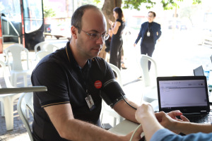 Foto do servidor Eduardo durante campanha. Trata-se de um homem branco calvo dos olhos claros; Ele veste uma camiseta preta e o crachá de identificação do TRT-18. Ele está sentado atrás de uma mesa branca, com o braço esquerdo estendido e dentro do medidor de pressão arterial.