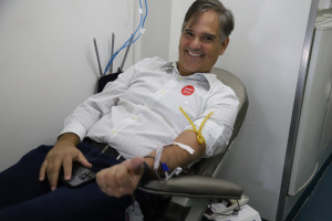 Foto do executivo comercial Maurício Botilho durante doação de sangue. Trata-se de um homem branco com cabelo curto, liso e grisalho. Ele veste uma calça social preta e blusa social branca. Na imagem, o homem está deitado numa cadeira almofadada, com o braço estendido e com acesso ligado à bolsa de sangue. 