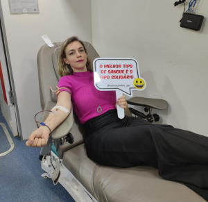 A mulher na imagem está doando sangue em um ambiente hospitalar ou clínico. Ela está deitada em uma cadeira de doação, com o braço estendido e conectado a um equipamento de coleta de sangue. Ela segura uma placa com a mensagem: "O melhor tipo de sangue é o tipo solidário", acompanhada de um emoji sorridente. Ela veste uma blusa rosa e calça preta, e parece tranquila durante o processo de doação.