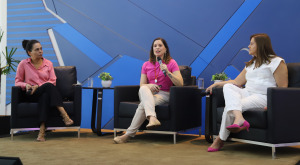 A imagem mostra três mulheres sentadas em poltronas, participando de uma conversa ou debate em um ambiente formal, possivelmente um evento ou painel de discussão. As duas mulheres nas extremidades parecem estar ouvindo enquanto a mulher ao centro fala, segurando um microfone. O fundo é decorado com um design geométrico em tons de azul, e há pequenas plantas decorativas sobre mesas entre as poltronas. As três mulheres estão vestidas de forma casual, mas elegante: uma usa rosa com preto, outra rosa com branco, e a terceira está vestida de branco com sapatos rosa.