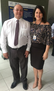 Um homem de camisa branca, gravata e calça escura ao lado de uma mulher de saia e blusa de manda sorriem pra foto