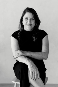 Mulher de cabelos médios, vestido preto, sentada e posando para foto com pernas cruzadas. A foto é Preto e branco.