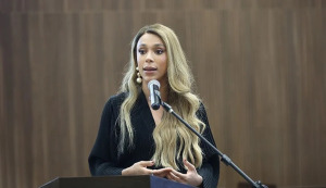 Mulher fala ao microfone. Ela tem cabelos longos e loiros e usa terno preto