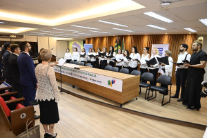 A imagem mostra um coral de pessoas, a maioria mulheres, vestindo blusas brancas e calças escuras, cantando em um ambiente formal, possivelmente um auditório. Elas estão em frente a uma mesa comprida com um banner que diz "CONGRESSO BRASILEIRO DE DIREITO E POLÍTICAS PÚBLICAS". À esquerda, algumas pessoas de terno estão de pé, de costas para o observador, observando o coral.