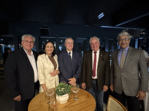 Quatro homens de terno e uma mulher também de traje social estão em pé, em frente a uma mesa pousando para foto.