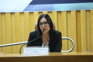 Foto da desembargadora Wanda Ramos, relatora do recurso. Trata-se de uma mulher branca com cabelo preto e liso na altura do ombro, ela veste uma blusa estampada preta e branca, um blazer preto e óculos preto. A mulher está sentada atrás de uma mesa de madeira e falando em um microfone de mesa fixo na mesma. Na sua frente, sore a mesa, há uma placa com seu nome e cargo escritos. 