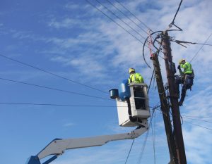 dois homens estão em cima de equipamentos para regular eletricidade em poste de energia