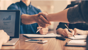 Duas pessoas dão as mãos celebrando acordo enquanto outra digita ao computador.