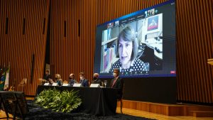 Foto mostra mesa diretora do evento com desembargadores sentados e um telão com a imagem da ministra do TST, Maria Cristina Peduzzi.