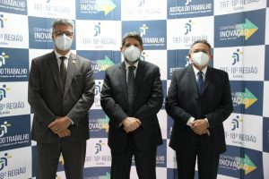 Presidente do TRT ao lado do Corregedor geral e do corregedor do TRTGoiás, todos de terno e gravata, em frente ao painel com logo do TRT Goiás