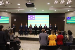 Auditório com pessoas e ao fundo a bandeira do Estado de Goiás