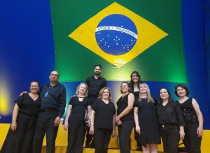 Foto colorida dos integrantes do Coral Labor em Canto com a bandeira do Brasil ao fundo