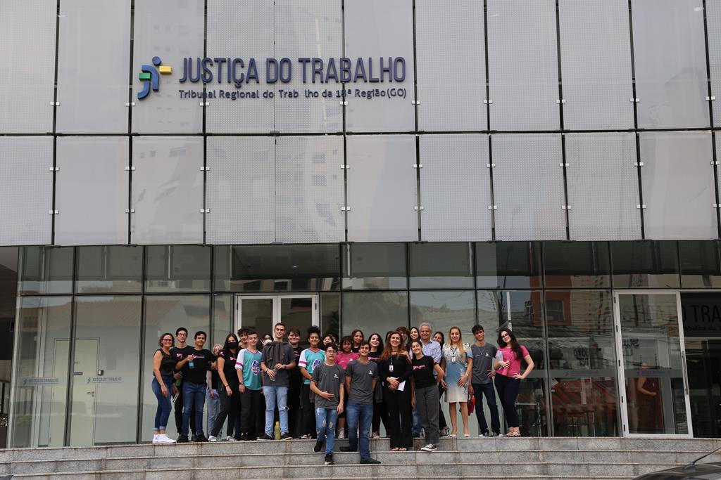 Grupo de 30 estudantes em frente à entrada do Tribunal, mostrando a fachada e o letreiro da JUSTIÇA DO TRABALHO ao fundo
