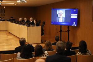 Na foto, vemos no plano de fundo os desembargadores presentes e, em destaque, televisão transmitindo a imagem do desembargador Eugênio. Em primeiro plano, as pessoas do público assistindo à sessão e à tv