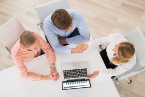 Três pessoas sentadas com um laptop e uma delas apresenta algo e aperta a mão da outra