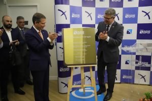 Dois homens de terno aplaudem a abeertura da agência com placa de inauguração