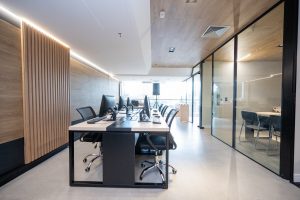 Sala bem iluminada com mobiliário novo, mesa ao centro com computadores e divisórias em vidro