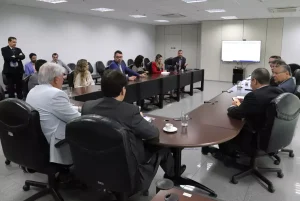 Foto dos participantes da reunião olhando para o telão da sala. 
