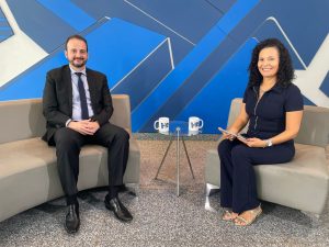 Duas pessoas sentadas, sendo o entrevistado advogado Paulo Henrique Pinheiro e a jornalista Fabíola Villela. No fundo, um painel azul e ao centro uma pequena mesa com duas canecas em cima