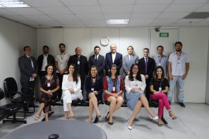 Imagem colorida de um grupo de dezesseis pessoas sorrindo para foto. Na frente, seis mulheres estão sentadas. Ao fundo, dez homens de pé.