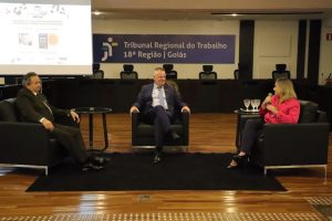 Dois homens de terno e uma mulher de camisa rosa sentados em sofás no palco em debate com a plateia