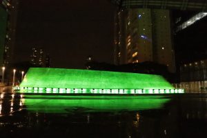 Foto noturna mostrando parte da estrutura do Complexo Trabalhista iluminado com a cor verde. Ao fundo, prédios com luzes acessas.