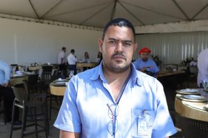 Homem com uma feição séria usando camisa de uniforme azul-claro. Um óculos de grau está pendurado na camisa. No bolso da camisa está preso um crachá de identificação.