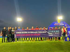 Foto do estádio de futebol com jogadores, alguns membros da delegação e dois magistrados em pé, lado a lado, seguram uma faixa de seis metros de largura com fundo roxo e os dizeres "Semana Nacional da Conciliação Trabalhista: você a um passo da solução! 22 a 26 de maio. Inscreva-se: csjt.jus.br", na faixa, também estão as logos da Conciliação Trabalhista e da Justiça do Trabalho.