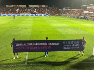 Na foto, três gandulas seguram a mesma faixa no campo de futebol durante intervalo, ao fundo, alguns jogadores em campo e a arquibancada cheia de torcedores.