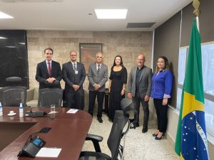 Seis pessoas em pé em uma sala com uma mesa de um lado e uma janela do outro
