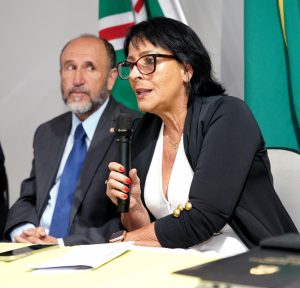 Duas pessoas, lado a lado, uma mulher e um homem. A mulher está com o microfone na mão falando