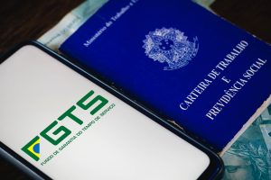 Foto colorida de um celular e uma carteira de trabalho colocados, lado a lado, sobre uma superfície plana. A tela do celular se encontra para cima e nela aparece uma imagem com a logo do FGTS.