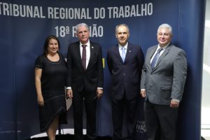 Quatro pessoas em pé, sendo uma mulher e três homens. Ao fundo um painel com as inscrições Tribunal Regional do Trabalho 18ª Região
