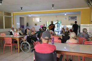 Foto de idosos durante programação musical no Solar Colombino. Os idosos estão espelhados, sentados, em uma sala repleta de mesas e cadeiras. No centro das mesas, um homem está de pé, cantando e tocando um violão.