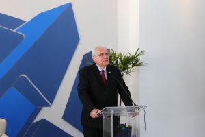 Foto do desembargador Wellington Peixoto discursando durante evento. Trata-se de um homem branco, de cabelo curto, liso e grisalho. Ele veste terno preto, gravata vermelha e óculos de armação preta. O homem está atrás de um púlpito e fala no microfone de mesa.