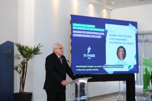 Foto do desembargador Wellington Peixoto discursando durante evento. Trata-se de um homem branco, de cabelo curto, liso e grisalho. Ele veste terno preto, gravata vermelha e óculos de armação preta. O homem está atrás de um púlpito e fala no microfone de mesa.