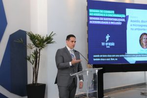 Foto de juiz Fernando Rossetto discursando durante evento. Trata-se de um homem branco, de cabelo moreno, liso e curto. Ele veste terno cinza e gravata roxa. O homem está atrás de um púlpito e fala no microfone de mesa.