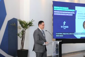 Foto de juiz Fernando Rossetto discursando durante evento. Trata-se de um homem branco, de cabelo moreno, liso e curto. Ele veste terno cinza e gravata roxa. O homem está atrás de um púlpito e fala no microfone de mesa.