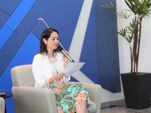 Foto de palestrante durante evento. A mulher está sentada em um poltrona branca, enquanto olha um papel em sua mão e fala no microfone de mão.