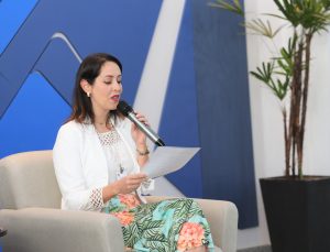 Foto de palestrante durante evento. A mulher está sentada em um poltrona branca, enquanto olha um papel em sua mão e fala no microfone de mão.