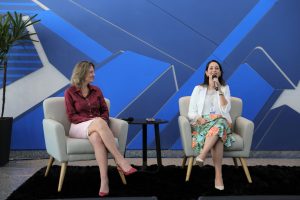 Foto de duas palestrantes durante evento. As duas estão sentadas lado a lado em poltronas separadas por uma mesinha de centro. Uma delas fala em um microfone de mão.
