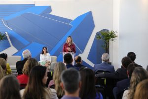 Foto de uma palestrante do evento, de pé. atrás do púlpito falando em um microfone de mesa. Na sua frente, a plateia a assiste.