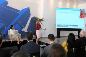 Foto de uma palestrante do evento, de pé. atrás do púlpito falando em um microfone de mesa. Na sua frente, a plateia a assiste.