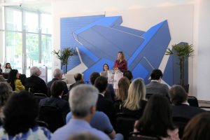 Foto de uma palestrante do evento, de pé. atrás do púlpito falando em um microfone de mesa. Na sua frente, a plateia a assiste.