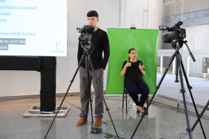 Foto do câmera e da intérprete de libras durante evento. O homem está de pé atrás da filmadora e a mulher sentada de frente para uma câmera para a qual faz os sinais de libra.