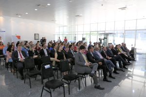 Foto de plateia durante evento. O público senta em cadeiras alinhadas verticalmente e horizontalmente na sala.