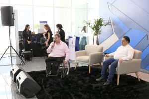 Foto de palestrantes durante evento. São dois homens. Um dos homens senta em uma poltrona. O outro está sentado na cadeira de rodas e fala em um microfone de mão.