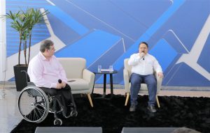Foto de palestrantes durante evento. São dois homens. Um dos homens senta em uma poltrona e fala em um microfone de mão. O outro está sentado na cadeira de rodas e segura um microfone no colo.